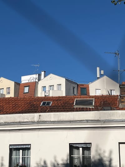 The image depicts the rooftops of buildings under a clear blue sky. A large, diagonal shadow cuts across the upper portion of the sky. Several buildings with terracotta tiled roofs are visible, with some featuring dormer windows. Various antennas and a chimney are present on the rooftops. The lower part of the image shows the facade of a white building with a large window that has a striped awning and sheer curtains. The scene appears to be from a residential area in the city of L'Haÿ-les-Roses, France.