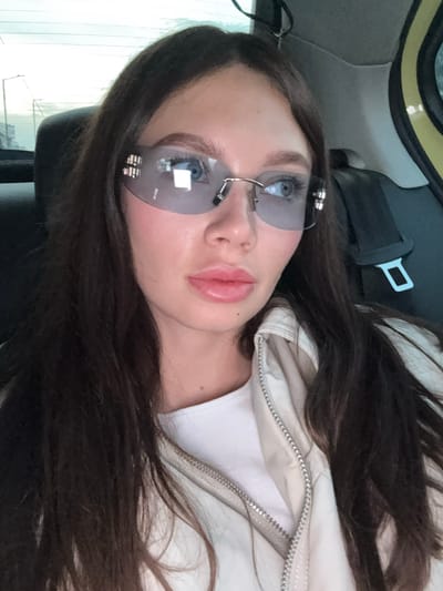 The image depicts a young woman with long, dark hair, wearing sunglasses and a light-colored jacket. She is inside a vehicle, likely a car, judging by the presence of a seatbelt and headrest. The location is possibly Agios Dimitrios, Greece, although this cannot be determined from the image. There are no actions or interactions apparent in the image. The environment appears to be the interior of a vehicle, with a glimpse of the exterior visible through the car window.