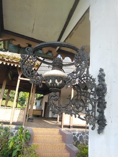 A large, ornate, black cast-iron wall-mounted lantern fixture is affixed to a white, weathered concrete pillar or wall in Dulce Nombre de Jesús, Costa Rica. The fixture, positioned prominently in the foreground, features intricate scrollwork surrounding a central opaque, light-colored glass globe with a textured, dark metal cap. Above the lantern, a light-colored soffit with peeling paint and dark brown trim is visible, supported by decorative wooden eaves in light brown and teal. Below the lantern, a set of three tiled steps, patterned in light brown squares, ascends towards a covered patio or veranda. This structure, partially visible in the midground, has dark wooden posts, a dark roof, and a visible concrete or tiled floor. To the left of the steps, green foliage and bushes are present, bordering a dark, rough-hewn stone base or wall. No individuals, actions, or specific interactions are depicted. The scene is illuminated by natural daylight.