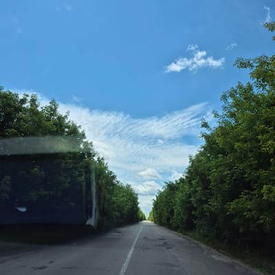An asphalt road stretches into the distance, flanked by dense green trees on both sides. The road has a white dashed line down the center, and patches of damage are visible on the surface. The sky above is a bright blue, with scattered white clouds. The image appears to be taken from inside a vehicle, as evidenced by reflections on a dark surface on the left side of the frame, likely a car window. The scene captures a rural or semi-rural setting in or near the city of Fursy, Ukraine.