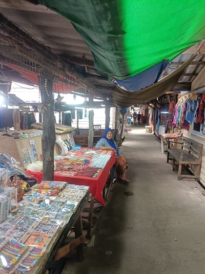 This media file captures a lively market scene in Tambon Ko Panyi, Thailand. A woman, wearing a blue hijab and an orange patterned skirt, sits behind a table laden with jewelry and trinkets. The table is draped with a vibrant red cloth, displaying an array of necklaces, bracelets, and other accessories. In front of her, another table is covered with various packaged items, possibly souvenirs or small goods.

The market stalls are covered with a patchwork of tarps, primarily green and blue, providing shade from the sun. The setting appears to be an open-air walkway, with wooden structures and benches lining the path. Clothing items, like colorful t-shirts and dresses, are hung on display on the right side of the walkway. Further down the path, a few figures can be seen walking, suggesting a gentle flow of activity.

The overall atmosphere seems relaxed and somewhat casual, with the woman displaying a calm demeanor. The lighting suggests it's daytime, possibly late morning or early afternoon, with sunlight filtering through the tarps. The concrete ground shows some wear and tear, typical of a well-trodden marketplace. The image provides a glimpse into the daily commerce and cultural fabric of this Thai location.