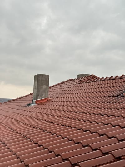 The image displays a terracotta-tiled roof in Baiachevo, Bulgaria, viewed from an elevated perspective under an overcast grey sky. The roof is composed of numerous parallel rows of reddish-brown, curved tiles. Two chimneys protrude from the roofline. The most prominent chimney, located on the left half of the frame, is a grey, rectangular concrete structure with visible metal flashing, including a red section, at its base where it interfaces with the roof tiles. A second, smaller grey chimney is partially visible further to the right. The tiles exhibit some minor surface variations and possible dampness or staining in certain areas, particularly towards the right side of the frame. A faint, dark landmass is visible on the distant horizon to the left, beneath the sky. No individuals, specific actions, or interactions are present in the scene.