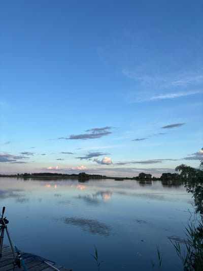 The image depicts a landscape at sunset, possibly near Khryashchevka, Russia. The scene includes a body of water, likely a lake or river, with reflections of the sky and trees. The sky features a mix of blue and various clouds illuminated by the setting sun. A treeline is visible along the horizon, its reflection mirrored in the water. In the foreground, there appears to be a wooden dock or platform with fishing equipment present, suggesting recreational fishing. Reeds and vegetation are visible along the water's edge. No people are visible in the image.
