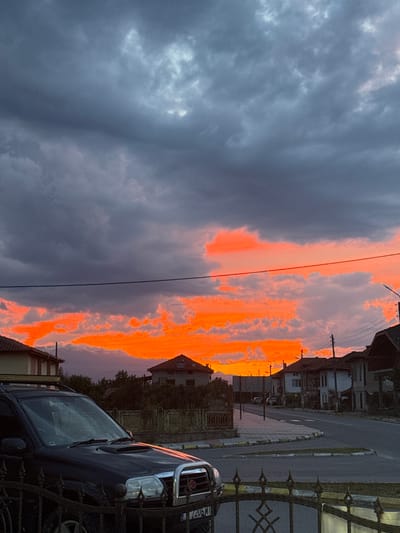 This image captures a dramatic sunset over a residential street in Blatska, Bulgaria. The sky is dominated by dark, heavy clouds in the upper portion, contrasting sharply with the vibrant, fiery orange and red hues of the lower sky, indicating the sun is setting behind the clouds. Scattered, lighter clouds are interspersed with the bright colors.
In the foreground, a dark-colored SUV is parked on the left side of the street, partially obscuring a decorative metal fence. The car's headlights and front grille are visible, along with part of its license plate which reads "7206 MT".
The street itself is paved, with a sidewalk and curbing visible. Several houses, typical of Bulgarian village architecture with tiled roofs, line the street. Some have visible windows and chimneys. A street lamp is visible on the right side of the image. In the distance, mountains are faintly visible, silhouetted against the colorful sky. The overall impression is one of a quiet, semi-rural setting experiencing a spectacular natural phenomenon. There are no people visible in the image.