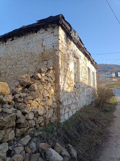 An old stone building with a crumbling wall and dilapidated roof stands adjacent to a dirt path. Two window openings are visible on the side. A stone wall, partially collapsed, sits at the corner of the building. Dry grasses and vegetation grow along the path. In the background, residential buildings are seen on a hillside under a clear blue sky.