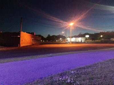 This nocturnal photograph depicts a quiet, deserted commercial area in Tahlequah, United States of America, illuminated by artificial lights. In the immediate foreground, a vibrant, bright purple-colored pathway stretches diagonally from left to right, bordered by a strip of dark grass with small white wildflowers.

The middle ground is dominated by a wide, empty asphalt surface, possibly a street or parking lot, which reflects some of the ambient light. To the left, a multi-story brick building with a ramp leading to an entrance is dimly lit. A prominent, intensely bright orange street light stands centrally, emitting strong lens flares that radiate across the dark sky, revealing faint, wispy clouds tinted reddish-orange.

To the right, another building, possibly a gas station or convenience store, is brightly lit from within, with its exterior and a canopy also illuminated. An unreadable sign stands in front of this building. Utility poles and overhead wires are visible against the backdrop of the sky. In the background, a line of dark trees marks the horizon, with other distant, smaller lights twinkling. The sky transitions from a deep dark blue overhead to a lighter blue-grey near the horizon. A faint crescent moon is visible in the upper right sky.

The scene is devoid of people or vehicles, conveying a sense of stillness and quiet. No discernible text is visible on any signs or structures within the image.