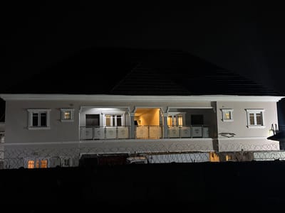 The image depicts a two-story residential building at night in Angwan Sayawa, Nigeria. The building features a light-colored (beige or off-white) facade with a dark, sloped roof. Multiple white-framed windows are visible across both floors.

A prominent balcony spans a significant portion of the upper floor, supported by columns. It is enclosed by glass railings with frosted decorative patterns. Several rooms within the house, particularly those opening onto the balcony and some upper-floor windows, are illuminated with warm-toned interior lights, suggesting occupancy.

In the foreground, a dense, spiraling razor wire fence runs horizontally, acting as a security barrier. This fence partially obscures the lower section of the building, though some illuminated windows are still visible below it. The sky above the house is completely dark, indicating a clear night. No people or visible text are present in the image. The scene portrays a well-lit home, secured by a formidable fence, under the cover of darkness.