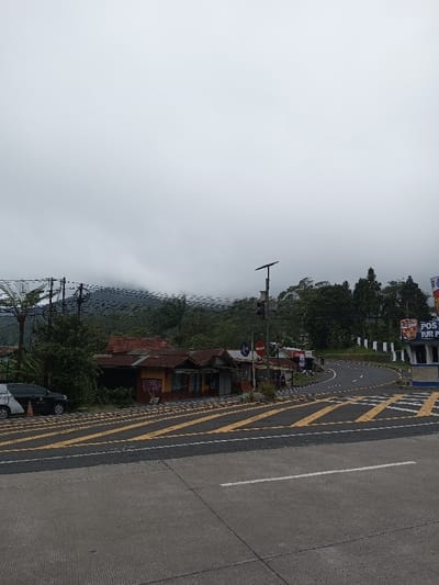 The image displays an outdoor scene in Phoxay, Laos, under an overcast sky with low-hanging clouds partially obscuring green mountains in the background. The foreground consists of a concrete road surface marked with a single white transverse line. In the midground, an asphalt road curves sharply to the right, featuring multiple yellow diagonal striped markings on its surface, indicating a designated turning lane or caution area. To the left of this road, a series of three single-story buildings with reddish-brown corrugated metal roofs and weathered facades (painted in shades of yellow and brown) are visible. A white sedan and a dark sport utility vehicle (SUV) are parked on a dirt shoulder beside these buildings, with an orange and white traffic cone positioned near the white sedan. A tall utility pole, fitted with a solar panel at its peak, stands near the roadside, supporting a dense network of overhead power lines that stretch across the mid-horizon. Adjacent to this pole is another pole bearing a red circular traffic sign. To the right side of the road, a blue and white structure, likely a small booth or guard post, is present, displaying a sign that partially reads "POS PUR P". Dense green vegetation and trees line the road and cover the distant hills. No individuals are clearly discernible in the scene.