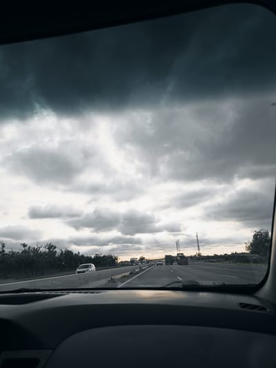 The image shows a view from inside a car driving on a highway. Outside, the sky is overcast with dark, heavy clouds. In the distance, there are electrical towers and vehicles ahead on the road. The landscape alongside the highway consists of low vegetation. The foreground is the dashboard of the car. The location is Shoul السهول, Morocco.
