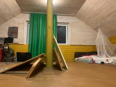 The image shows an attic room in a house located in the village of Барское-Городище, Russia. The room has sloped wooden ceilings and walls. In the center of the room, a large wooden pillar supports the ceiling. Two green curtains are visible, one behind the pillar and another to the left. There are two windows in the room; one is to the left of the pillar, partially covered by the green curtain, and another is to the right. In the foreground, two large, folded wooden panels are propped up against the pillar. To the left, a black suitcase and a large, dark, sack-like object are placed on a patterned rug. Further to the left, a framed picture hangs on the wall. In the background, on the right side of the room, a bed with a mattress and white bedding with floral patterns is visible, along with a black headboard. A white mosquito net is suspended from the ceiling over the bed. The floor appears to be made of polished wood. The overall impression is of a functional, somewhat unfinished living space.