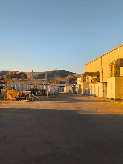 The image depicts an industrial area bathed in the warm glow of what appears to be late afternoon or early morning sun. In the foreground, a paved lot leads towards a row of low-lying industrial buildings. These buildings are primarily white and tan, with some featuring large, external ventilation ducts that are a reddish-brown color, suggesting age or rust. To the right, a larger, yellow-sided industrial building dominates the scene. Piles of wooden pallets and yellow plastic crates are stacked near the buildings on the left side of the lot. Beyond the structures, a gently sloping, arid landscape is visible under a clear, pale blue sky. A few trees are scattered on the hillsides, and a tall, slender structure, possibly a communication tower or smokestack, is visible on the crest of the furthest hill, topped with a yellow beacon or light. There are no people or vehicles visible in the image. The overall impression is one of a functional, perhaps slightly aged, industrial complex in a dry, open environment.