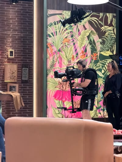 The image shows a studio setup in Sant Joan Despí, Spain, possibly for a television production. In the foreground, a large, plush, light brown sofa occupies the bottom half of the frame. Behind it, a man wearing a black t-shirt, black shorts with white stripes, and headphones is operating a camera stabilizer rig, which supports a professional video camera. He is facing away from the viewer, towards a vibrant backdrop featuring a tropical foliage pattern with pink and green tones. To the right of the man, a woman wearing a black top and headphones is also present, seemingly observing or participating in the production. She is looking towards the right side of the frame. The background wall to the left is made of exposed brick, with several framed pictures and a narrow vertical light strip. Overhead, a lighting fixture with a camera attached to it is visible. The overall impression is that of an active filming environment.
