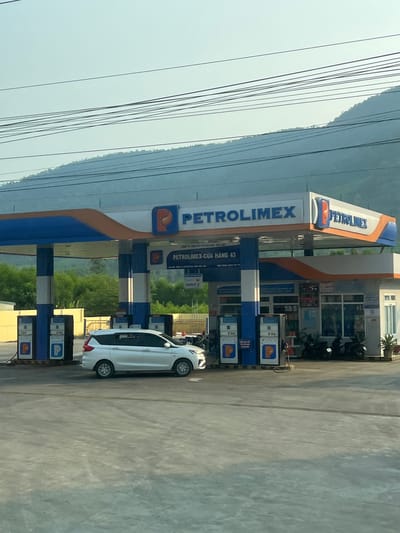 Here's a detailed description of the image:

**Setting:** The image depicts a Petrolimex gas station.

**Specifics:** The Petrolimex station has multiple fuel pumps, and a white car is parked near one of them. The station also appears to have a convenience store and office area. Mountains are visible in the background. Utility lines run across the upper left portion of the image.

**Text on the Station:** Prominently displayed is the brand name "Petrolimex". The sign below the logo says "PETROLIMEX-CỬA HÀNG 43," which translates to Petrolimex-Store 43. 

**Location:** The station is located in Thôn 18, Vietnam.