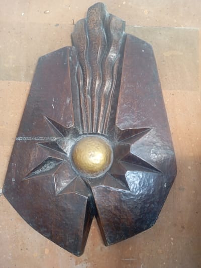 This is a close-up, overhead shot of a bronze sculpture mounted on a textured, light brown wall, likely made of stone or tile. The sculpture is abstract and geometric, featuring a central golden sphere surrounded by sharp, star-like points. Above the sphere, vertical, wavy lines suggest flames or flowing elements. The entire piece has a rough, hammered texture, indicating it's a crafted object. A thin, jagged line, possibly a weld or inscription, is visible around the star points. The overall impression is of a sun or a radiant symbol, possibly representing an important cultural or religious motif. The setting appears to be indoors, given the lighting and the uniformity of the background. There is no visible text. The location is identified as Ararat, Armenia.
