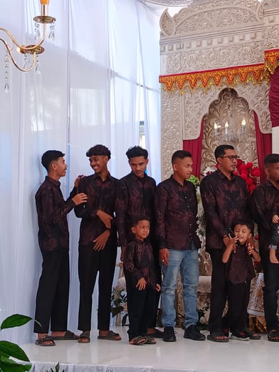 A group of eight individuals, six adult males and two young boys, are standing in a line on a raised platform in front of white curtains. They are all wearing matching dark patterned shirts and dark pants. The adult males are of varying ages and ethnicities, and all appear to be of Indonesian descent. The two young boys are positioned in the front row, one of whom is holding a mobile phone. The background features a decorative backdrop with intricate carvings and red and gold trim, suggesting a formal event or celebration. A chandelier hangs from the ceiling to the left of the frame, and a large floral arrangement is visible behind the group. The platform they are standing on is white and appears to be covered with a white cloth. The location is identified as the city of Meuliek, Indonesia.
