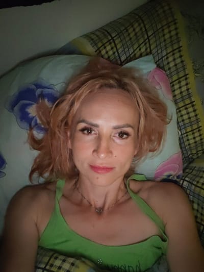 A woman with light brown, shoulder-length hair is lying on a pillow in bed. She is looking directly at the camera. She is wearing a green tank top. The pillow has a floral pattern with blue and pink flowers. The background shows a plaid blanket. The lighting appears to be artificial. The image was taken in Shalsky, Russia.