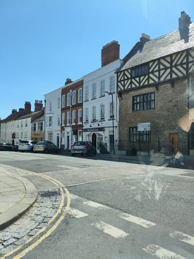 The image shows a street scene in Ludlow, United Kingdom, on a sunny day. The view is from a vehicle, with reflections visible on the windshield. On the right side of the image, a building with a stone lower facade and timber-framed upper stories is prominent. This building has a dark tiled roof and a wooden door. To its left, a row of attached buildings with white and red brick facades extends down the street. These buildings have multiple stories with numerous windows. Several cars are parked along the street. In the foreground, a road with white pedestrian crossing lines and double yellow lines is visible. A curved paved area with cobblestones and a drain cover is on the left. The sky is clear and blue.