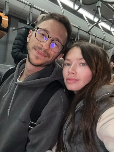 The image displays two individuals, an adult male and an adult female, captured in a close-up selfie within the interior of a public transport vehicle, consistent with a metro train on the Athens Metro system, which serves the Paiania Municipal Unit, Greece. The male, positioned on the left, has short brown hair, a short beard, and wears black-framed glasses, a grey hooded sweatshirt, and a black backpack strap. He presents a slight smile while looking directly at the camera. The female, on the right, possesses long dark brown hair, light-colored eyes, and wears a grey zippered jacket over a light-colored garment. Her gaze is directed towards the camera with a neutral expression. The overhead environment features numerous grey strap handles attached to metal bars and a horizontal fluorescent light fixture. A blurred individual in a dark puffer jacket is partially visible behind the male, and another obscured figure is in the distant background on the right. An orange light source is visible through a window in the upper left, indicating an external environment, likely at dusk or night.