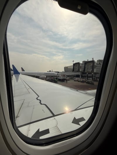 An airplane window frames the view of a wing and the terminal. The wing, painted white, features black "NO STEP" markings and arrows. A Copa Airlines plane sits at the terminal, with support vehicles clustered around it. The sky is visible above the terminal, filled with clouds. The window's frame is black and the window shade is partially drawn.