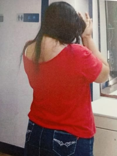 The image captures a person from behind, standing indoors, likely in Kaohsiung, Taiwan. The individual has dark, shoulder-length hair and is wearing a bright red short-sleeved top that is off-the-shoulder on the left side, revealing a white strap underneath. They are also wearing dark blue jeans with intricate white embroidery on the back pockets.

The person's right arm is raised, with their hand near their head, suggesting they might be adjusting their hair or performing some personal grooming. To the left, on a light-colored wall, a dark blue sign is visible. This sign features universal male and female restroom symbols, and to their right, two visible Chinese characters. These characters are "廁所" (cèsuǒ), which translates to "restroom" or "toilet," strongly indicating the setting is a public washroom. On the right side of the frame, a reflective surface, possibly a mirror or window, and a light-colored counter or ledge are present. The overall setting is an indoor, public space.