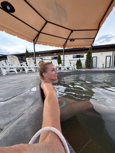 A middle-aged Caucasian woman is relaxing in a pool, her shoulders and neck above the water. She is looking towards the upper left, her blonde hair pulled back in a bun, wearing hoop earrings. She is resting her arm on the edge of the pool, with a light-colored bracelet on her wrist. The pool's edge is made of a grey concrete material. White plastic lounge chairs are lined up behind her. In the background, there is a multi-story building. A beige canopy covers part of the pool area. The setting is Kara-Oy, Kyrgyzstan.