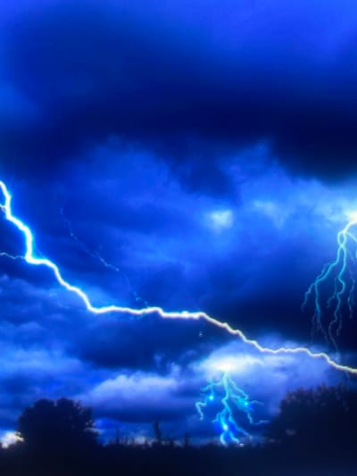 The image captures a stormy, nocturnal scene over Zelenyi Hai, Ukraine. The sky is dominated by deep blues and dark grays, indicative of heavy cloud cover during a thunderstorm. Multiple lightning strikes are visible across the frame. A prominent, bright white to electric blue lightning bolt extends from the upper left corner, diagonally across the middle of the image, curving towards the bottom-center. On the far right, a branched, electric blue lightning strike illuminates the clouds. A third, similarly branched electric blue bolt is present near the bottom-center, appearing to strike the ground. The lower third of the image features a dark, silhouetted horizon line, composed of dense foliage and trees against the illuminated sky. No human subjects or discernible man-made structures are present.