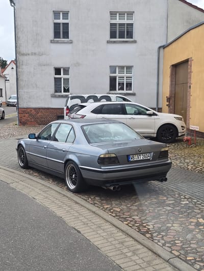 This image depicts a street scene in Kemberg, Germany. In the foreground, a gray BMW sedan is parked on a cobblestone street, with its exhaust emitting a visible plume of smoke. The car has a license plate that reads "WB AT 2604". Behind the BMW, a white SUV is parked, and further back, a white van with a black graphic is visible. The background features a light-colored building with multiple windows, and a yellow building with a wooden garage door. A red fire hydrant is also present near the yellow building, along with a red and white sign that reads "Saugstelle". The sky is overcast.
