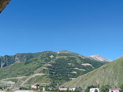 The image depicts a panoramic view of a mountainous landscape with a town nestled in the valley below, likely Tyrnyauz, Russia. The sky is a clear, vibrant blue. In the foreground, a portion of a textured, rough surface, possibly concrete or stone, juts into the frame from the top left corner.
The middle ground is dominated by rolling green hills and mountainsides, extensively terraced and dotted with dark green trees, suggesting agricultural or reforestation efforts. Several winding roads or paths are visible carved into the slopes. Power lines and transmission towers are scattered across the terrain, indicating infrastructure.
At the base of the mountains, a town is visible. Buildings of various sizes and architectural styles are present, including what appears to be a partially constructed or abandoned building with a grid-like facade. A tall, red and white radio tower stands out among the structures. Further into the valley, more buildings are clustered together, some with red roofs.
The background features larger, rugged mountains, some with lighter-colored rock faces and snow-capped peaks, emphasizing the alpine environment. The overall impression is one of a settlement in a remote, mountainous region. A timestamp in the bottom right corner reads "02.07.2026 09:10". No people are discernible in the image.