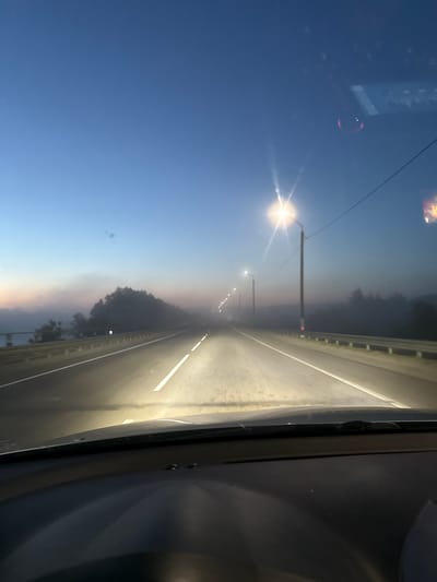 The image depicts an evening view from inside a vehicle traveling on a bridge or elevated road, likely in Krychau, Belarus. Streetlights illuminate the road, with their light appearing to scatter in a slight haze. The sky transitions from a pale blue to a pinkish hue near the horizon. Guardrails flank the road. The interior of the vehicle is partially visible on the right side of the frame.
