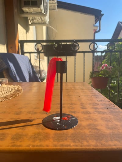 A red candle, bent and partially melted, stands in a black candle holder on a brown table. The table is set on a balcony with a black metal railing. In the background, a cream-colored building with a dark roof is visible, along with greenery and a blue sky. The scene appears to be outdoors, likely on a sunny day. The location is identified as Sremska Kamenica, Serbia.