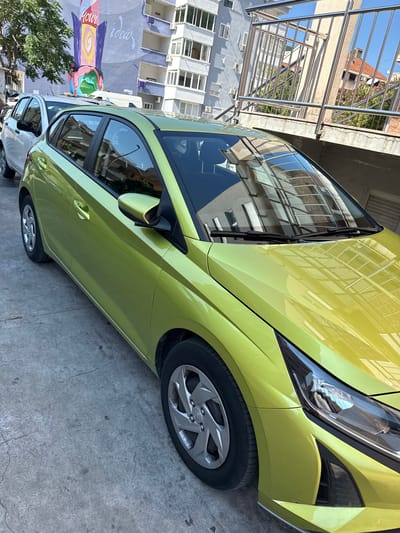 The image captures a bright, possibly sunny day in Lapčići, Montenegro, with a lime green car parked on a concrete surface. The car is the primary subject, angled from the front passenger side, showcasing its sleek design and reflective surface. The car's bodywork gleams, mirroring the surrounding buildings and a metal railing structure.

Behind the car, a multi-story building with numerous windows and balconies is visible. To the left, a large mural adorns the lower part of another building, featuring a colorful depiction of a rocket with planets and abstract shapes, and the words "Your ideas" written on it. A large, leafy green tree partially obscures the mural and the building. The overall scene suggests an urban or semi-urban environment with residential buildings and street art. The clear blue sky and the bright lighting indicate daytime, likely morning or afternoon. No people are visible in the image, and the car appears to be parked, with no activity taking place. The concrete ground shows some minor cracks and stains, suggesting it's an outdoor parking area or a public space. The metal railing in the background, likely part of a building's exterior or a walkway, adds to the architectural context of the location.