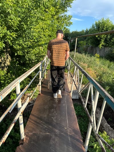 A man is walking away from the camera on a narrow metal bridge. He is wearing a patterned brown shirt, black pants, and white sneakers. He also has a black baseball cap on. The bridge has a rusted metal railing. The surrounding environment is overgrown with green vegetation and trees. There is a blue sky with some clouds visible. The scene appears to be in an outdoor, possibly rural or semi-rural, setting.