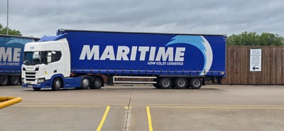 An outdoor, medium shot captures a white and blue Scania semi-truck and trailer parked in a lot. The trailer is predominantly blue with large white lettering spelling "MARITIME" and the words "LOW CO₂ST LOGISTICS" below it. The truck cab is white with blue accents. The truck is parked on a gray asphalt surface with yellow painted lines. In the background, a wooden fence with a white sign is visible. The sign has black text and a black arrow pointing to the left. Trees are visible behind the fence. The sky is overcast.