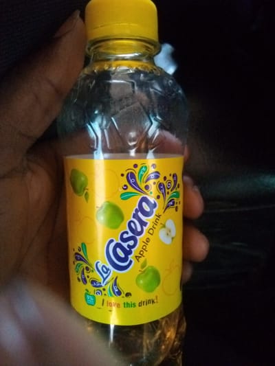 A person's hand is holding a clear plastic bottle of "La Casera" Apple Drink. The bottle has a bright yellow label with green, purple, and blue decorative swirls. The text "La Casera" is prominently displayed in a stylized font. Below this, the words "Apple Drink" are written, accompanied by an illustration of a sliced apple and several green apples. Near the bottom of the label, the phrase "I love this drink!" is visible. A small green circle with "5%" and "APPLE" printed inside is also present on the label. The background is dark, suggesting the photograph was taken indoors or at night, with no discernible weather or specific location cues beyond the provided context of Bokkos, Nigeria. The lighting focuses on the bottle, highlighting its contents and labeling.