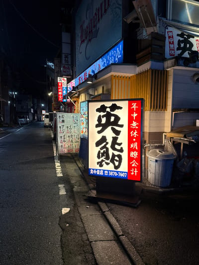 A nighttime street scene in Adachi, Japan features a brightly lit restaurant sign on the right, displaying Japanese characters and a phone number: 3870-7607. The sign is predominantly white with large black characters on the left, and red text on the right. To the left of the main sign, there are other illuminated signs and a sidewalk lined with more upright signs. A white trash bin is visible next to the main sign. The street itself is asphalt with white markings and is empty of pedestrians. Several parked vehicles, including a white van, are visible further down the street. Buildings with illuminated windows line the left side of the street. The overall atmosphere is that of a quiet urban street at night, with a focus on the glowing signage of a local establishment.