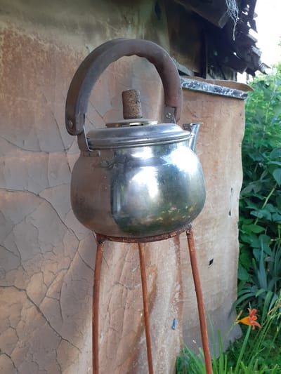 In Deremezna, Ukraine, a tarnished, metallic kettle sits atop a rusty, three-legged stand in front of a textured, weathered wall. The wall, adorned with an abstract, cracked pattern, serves as a backdrop to the scene. To the right, lush green foliage, including a vibrant orange lily, adds a touch of nature's color to the otherwise rustic setting. The overhead eaves of a structure are visible at the top of the image, suggesting a possible outdoor or semi-outdoor location. The scene is captured in daylight, with soft lighting illuminating the kettle and its surroundings.
