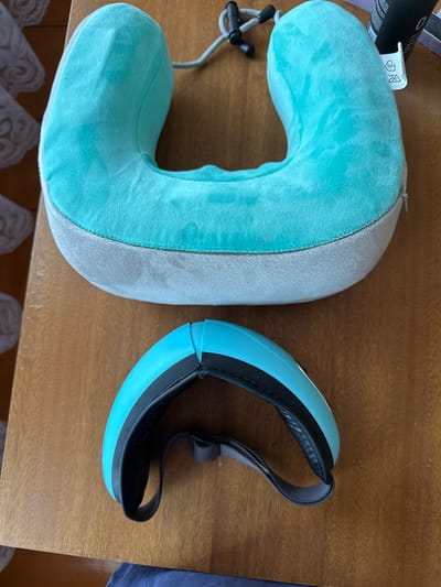 An overhead shot shows a turquoise neck pillow and a turquoise eye mask on a wooden table. The neck pillow is a U-shaped cushion covered in a soft fabric, with a grey lower section. A small zipper is visible on the right side of the pillow. Attached to the neck pillow are two black plastic clasps and a grey cord. Below the neck pillow, the turquoise eye mask is positioned horizontally. It has black padding around the edges and a grey adjustable strap. The wooden table has a visible grain pattern. In the background, to the left, a white lacy fabric is partially visible. To the right of the neck pillow, a black bottle with a white label and some other objects are partially in view. The scene is indoors, suggesting a personal or domestic setting. There is no indication of the city of Urazovo, Russia in the image.