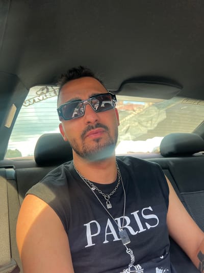 A man is sitting in the back seat of a car. He is wearing sunglasses and a black T-shirt with the word "PARIS" printed on it in large white letters. He is also wearing two silver chains around his neck. The car's interior is visible, including the headrests and seat upholstery. The view through the car window shows a blurred outdoor scene. There is no information indicating this scene takes place in Касапите, Bulgaria.