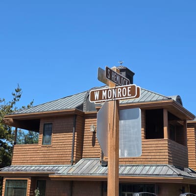 In Cannon Beach, Oregon, United States of America, a two-story wooden house with a shingled exterior is shown on a clear, blue sky day. A prominent wooden pole stands in front of the house, supporting two brown street signs with white lettering. The upper sign reads "S HEMLOCK" and the lower sign reads "W MONROE." To the left of the pole, a portion of the house's roof is visible, which appears to be made of dark gray metal. A small, decorative wooden fish carving protrudes from the eaves of the roof on the left side of the image. A weather vane is visible on top of a dark cupola on the roof in the background. The house has several windows with dark green frames. Trees with green foliage are present on the left side of the image.