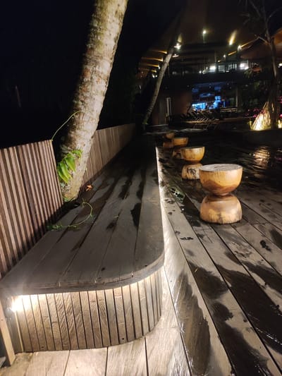 The image displays an illuminated outdoor wooden deck area at night in Mas, Indonesia. The dark-stained wooden deck exhibits numerous wet patches and streaks. On the left, a thick, light-colored tree trunk, possibly a palm, rises vertically, featuring green vine-like foliage attached to its bark. Adjacent to the tree, a curved, vertical-slat wooden structure functions as a bench, with a continuous light strip integrated along its base. Five distinct, hour-glass shaped wooden stools or low tables are arranged in a linear progression on the deck, extending from the mid-foreground towards the background on the right. In the background, a multi-level, modern building with extensive glass windows and dark horizontal architectural elements is brightly lit from within, revealing interior spaces. Several dark lounge chairs are positioned in front of the building, indicative of an outdoor seating or poolside area. Additional trees and illuminated landscaping elements are visible surrounding the building. No individuals or specific actions are present in the scene.