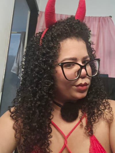 A woman with dark curly hair, wearing glasses and red devil horns, poses in front of a curtain and a dark background. She is wearing a red top with straps. The scene does not provide sufficient detail to identify a specific location within the urban perimeter of Cúcuta, Colombia.