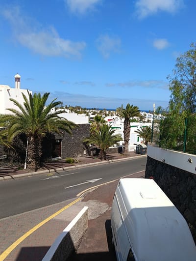 This sunlit outdoor image captures a street scene in Tías, Spain, characteristic of a residential or resort area. The time appears to be mid-day, with bright sunlight casting clear shadows under a vibrant blue sky dotted with scattered white cumulus clouds, indicating warm and clear weather.

The scene features numerous white, low-rise buildings, many incorporating dark, volcanic-looking stone into their bases or walls. A distinctive building on the far left has an ornamental, minaret-like tower with red and white detailing. Abundant palm trees are scattered throughout the landscape, with a large, prominent one on the left and others extending into the mid-ground, contributing to a subtropical ambiance. Some buildings in the distance display green or red window shutters.

An asphalt road curves through the center, marked with white directional arrows and a yellow painted line along the edge of the reddish-brown paver sidewalk. In the bottom right foreground, the roof and rear section of a white van are visible, parked next to a dark stone retaining wall. In the distance, more white buildings extend towards the horizon, where the clear blue ocean is visible. A grey public bin is discernible near the road in the mid-ground. There are no people visible, and no legible text can be found in the image. The overall impression is one of peacefulness in a characteristic Mediterranean coastal setting.