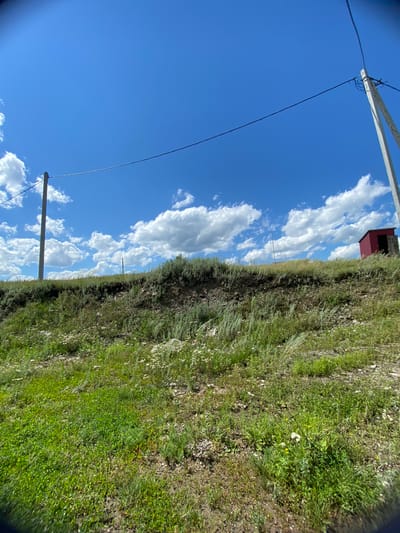 The image depicts an outdoor scene under a bright blue sky with scattered white clouds. In the foreground, a grassy slope with various green plants and some small white wildflowers dominates the lower half of the frame. A dirt path or disturbed earth is visible in the lower right portion. Two concrete utility poles with electrical wires stretched between them are present. The pole on the left is further in the background, while the one on the right is closer to the foreground and has wires connecting to it. On the top of the grassy ridge, a small, red, shed-like structure with an open doorway is situated. The environment appears to be a rural or undeveloped area.
