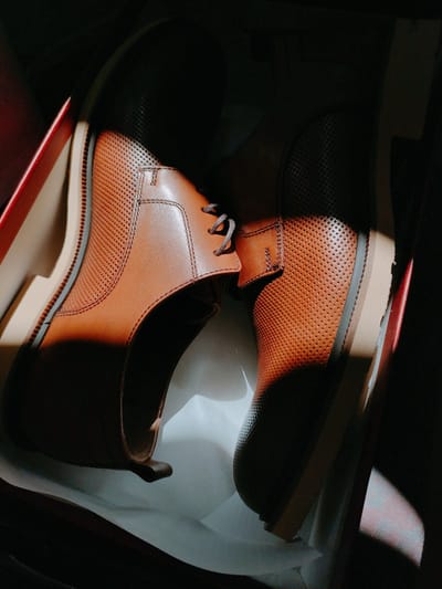 The image shows a pair of brown leather oxford shoes with perforated detailing, placed inside a shoebox. The shoes are positioned such that the light from a window casts dramatic shadows across them and the interior of the box. The shoebox itself appears to be made of cardboard with red accents on the lid's edge. The shoes are a light to medium brown color, with visible stitching and laces. The soles are a light tan color. The background is dark, suggesting the photo was taken indoors in a low-light environment, likely a home or retail setting. There is no information in the image to suggest any connection to Murska Sobota, Slovenia.