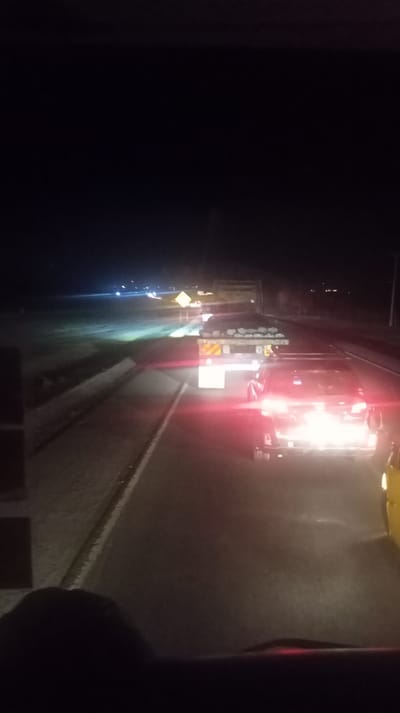 Night highway scene. A flatbed truck carries cylindrical objects, followed closely by a car with bright red taillights. A yellow vehicle is partially visible on the right. A diamond-shaped sign is noticeable on the left shoulder of the road. Other vehicles appear in the distance, illuminated by headlights. The view is from the driver's seat of a car.