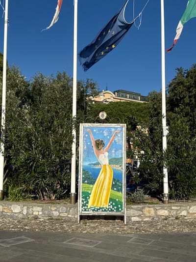 An outdoor daytime scene in Santa Margherita Ligure, Italy, is centered on a vertical, A-frame sidewalk sign. The sign features a painting of a woman with her arms raised, wearing a white strapless top and a flowing yellow skirt. Her face is obscured by green leaves. The background of the painting depicts a blue sea, a distant coastal town, green hills, and a foreground of grass with white daisies.
Behind the sign, three white flagpoles stand against a clear blue sky. A tattered blue European Union flag with yellow stars is visible on the central pole. A partially visible white and red flag is on the left pole, and a partially visible green, white, and red Italian flag is on the right pole. Dense green bushes and trees form a lush backdrop behind a low stone wall. Above the foliage, a large, ornate building with a light-colored facade and a dark roof is visible in the distance. The ground directly beneath the sign is cobblestone, transitioning to a darker, smoother pavement with rectangular inlays in the foreground.