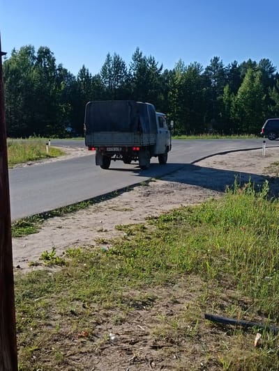 A light-colored truck with a dark tarp covering its cargo bed drives away from the camera on an asphalt road. The truck has a license plate that reads "M476TX 42". The truck is on a road that curves to the left, with a grassy shoulder and a dirt area to the right. In the background, there is a dense forest of green trees. The sky is a clear, bright blue, indicating it is daytime with fair weather. A black SUV is parked on the right side of the road in the distance. The scene appears to be in a rural or semi-rural area, given the presence of the forest and the relatively unpaved road shoulder.
