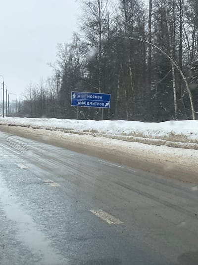 The image depicts a winter road scene from the perspective of a vehicle, looking forward. The road surface is dark, wet, and appears slushy or icy in patches, showing visible tire tracks and faint, worn white lane markings. Dirty, brown-tinged snow banks line both sides of the road, with a more substantial accumulation on the right. Beyond the snow banks, a dense line of bare deciduous trees, primarily birches, extends into the background, indicating a forested area. The sky is overcast and uniformly light gray.
On the right side of the road, a blue rectangular road sign is mounted on a pole. The sign contains two white panels:
1. The upper panel displays "A108 МОСКВА" (A108 MOSCOW) with an upward-pointing arrow, indicating that continuing straight on the A108 leads towards Moscow.
2. The lower panel displays "A108 ДМИТРОВ" (A108 DMITROV) with a rightward-pointing arrow, indicating that a right turn leads onto the A108 towards Dmitrov.
Utility poles with streetlights are visible receding into the distance along the left side of the road. No individuals or active interactions are visible in the scene.
The location is on the A108 (Moscow Big Ring Road), specifically at a junction or fork in the road near the village of Ivashevo, located in the Dmitrovsky District of Moscow Oblast, Russia. The sign directs traffic on the A108, with one path designated for Moscow (straight ahead) and another for Dmitrov (right turn).