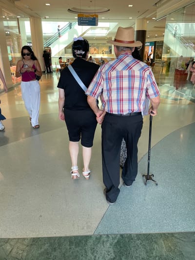 The media file shows a low-angle shot of four people inside a shopping mall. The primary subjects are two adults, likely a couple, standing in the foreground. The man, on the left, wears a plaid shirt, a hat, and dark pants, and is leaning on a cane. The woman, on the right, is wearing a black top and shorts, with white sandals. Behind them, another woman in a pink top and white pants is visible, holding a phone, and further in the background, a woman with black hair and a black top is seen.

The scene is set in a brightly lit shopping mall with a tiled floor and large glass windows revealing shops and escalators. Signs in Spanish are visible, indicating different levels and types of establishments within the mall, such as "supermercado" and "restauración." One sign reads "ARK" and another mentions "Cargado de posibilidades." The people appear to be casually strolling through the mall, suggesting a typical daytime shopping or leisure activity. There are no strong indicators of the time of day or weather, beyond the indoor lighting. The overall mood appears relaxed and ordinary, as expected in a public commercial space.