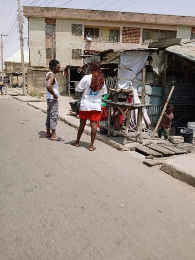 The image portrays a street scene in Mumbwa, Zambia, under clear daylight conditions. An asphalt road, displaying patches of wear, occupies the foreground, bordered on the right by a damaged concrete curb. Three individuals are present on the right side of the road, adjacent to a makeshift commercial structure. The subject on the left is an adult male with dreadlocks, wearing a white sleeveless top, grey cargo shorts, and sandals; he stands with hands on hips, oriented towards the right. Centrally positioned, an adult female with reddish-brown dreadlocks is observed from behind, wearing a white t-shirt with a blue "15" on the back and a red skirt; she is barefoot and faces the makeshift structure. A young child, partially visible on the far right, wears a striped top and holds a wooden stick, also oriented towards the right near the structure. This makeshift structure is assembled from corrugated metal sheets, wooden supports, and white fabric, featuring a wooden table, black buckets, and a green crate at its base, with a long wooden pole leaning against it. In the background, a multi-story building with a light-colored facade, balconies, and windows is visible, along with utility poles and overhead wires on the left.